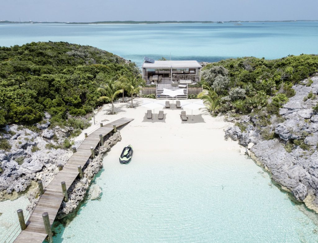 Staniel Cay, Exuma Bahamas A secluded beach house with a dock, surrounded by lush greenery and clear turquoise water, features a sandy area with lounge chairs and a small boat.