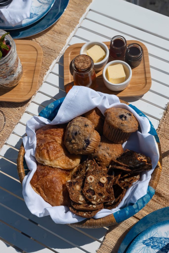 Staniel Cay, Exuma Bahamas A basket filled with muffins, croissants, and crackers on a table, accompanied by butter and small jars of jam.