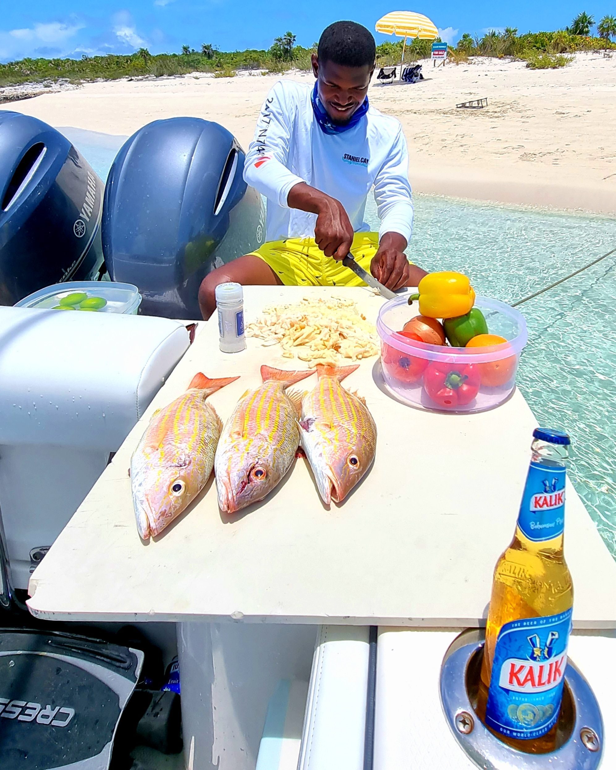 Staniel Cay, Exuma Bahamas Person cleaning fish on a cutting board on a boat by the beach. Three fish are laid out, with a bowl of vegetables and a Kalik beer bottle nearby.