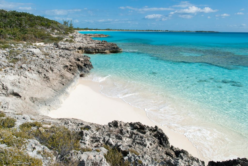 Staniel Cay, Exuma Bahamas Rocky coastline with clear turquoise ocean and a sandy beach under a blue sky. Sparse vegetation in the background.