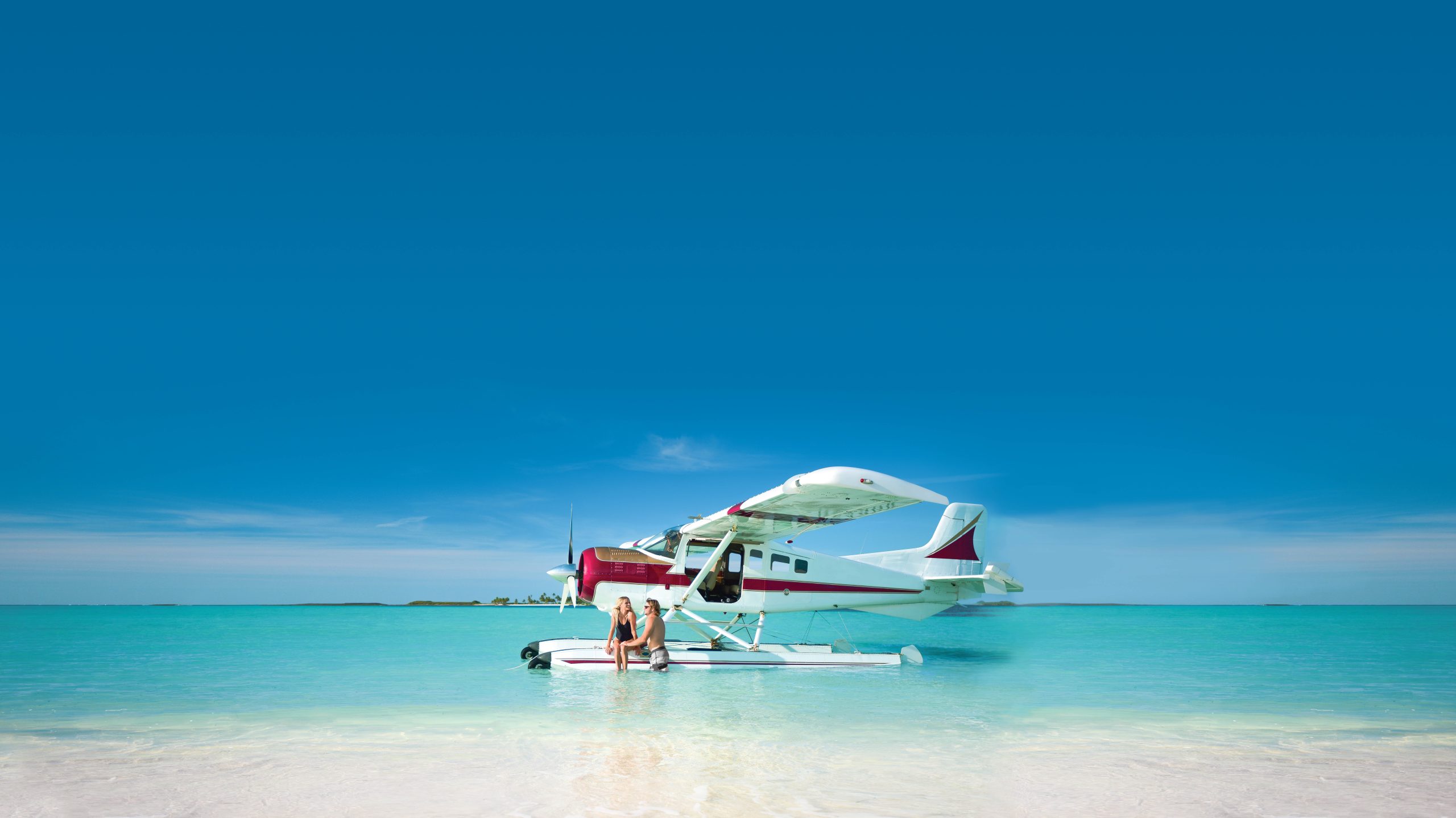 Staniel Cay, Exuma Bahamas A seaplane is anchored near a sandy beach with clear turquoise water under a blue sky. A person is sitting on its float.