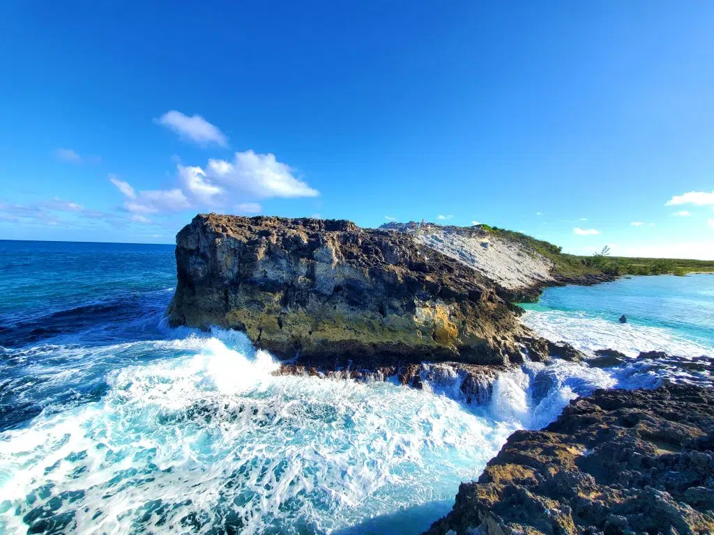 Staniel Cay, Exuma Bahamas Waves crash against a rocky cliff under a clear blue sky, with the ocean extending to the horizon.