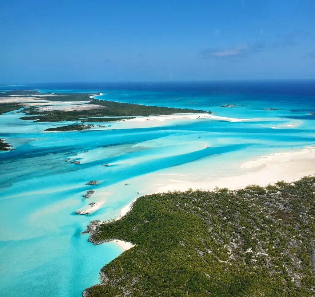 Staniel Cay, Exuma Bahamas Aerial view of a tropical coastline with turquoise waters, white sand beaches, and lush greenery under a clear blue sky.