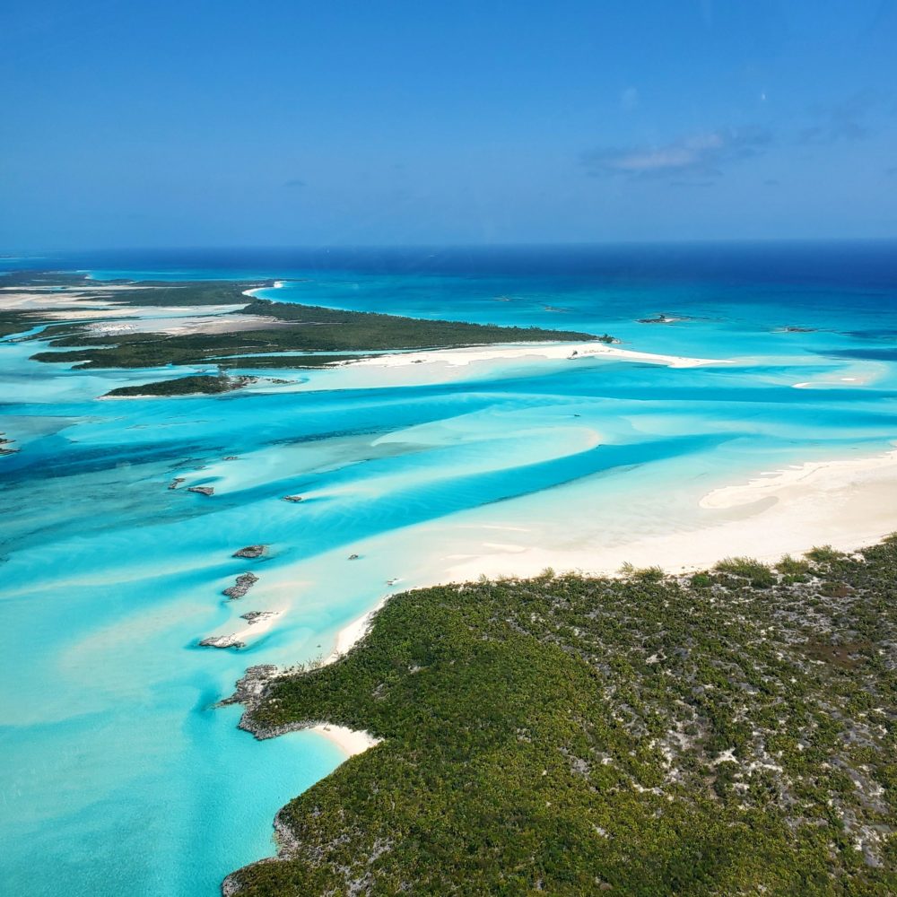 Staniel Cay, Exuma Bahamas Aerial view of a tropical coastline with turquoise waters, white sandy beaches, and scattered green vegetation under a clear blue sky.