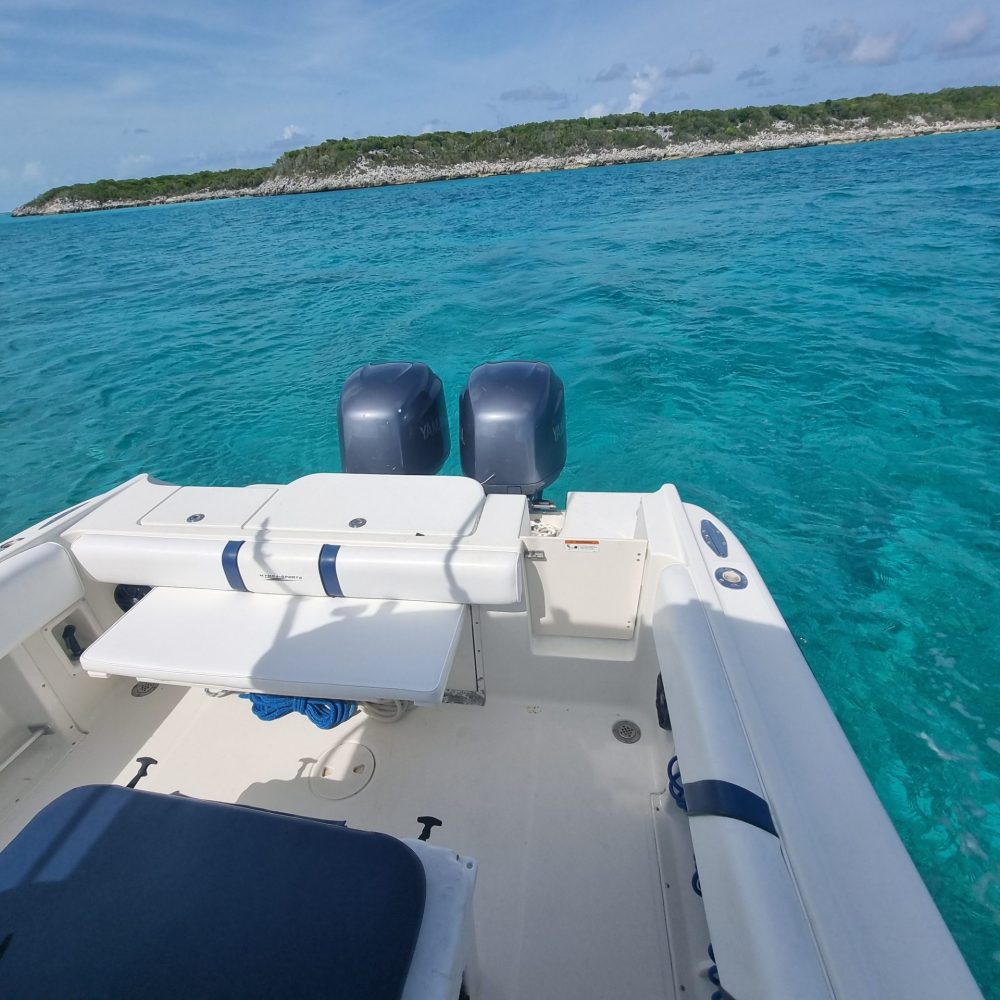 Staniel Cay, Exuma Bahamas View from a boat with white seating and dual outboard motors, facing clear turquoise water and an island with dense greenery under a blue sky with scattered clouds.