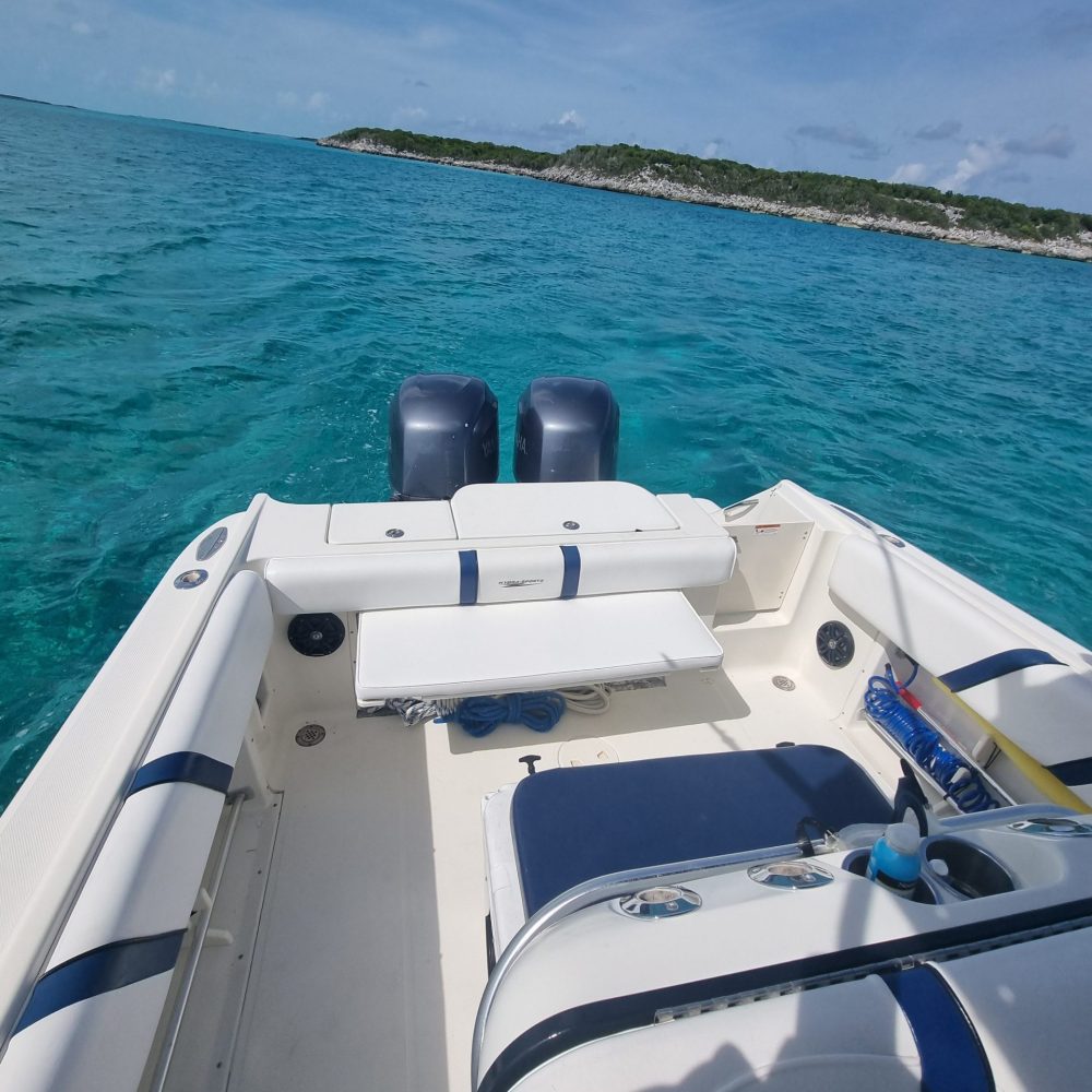 Staniel Cay, Exuma Bahamas The image shows the back of a boat with two engines, surrounded by clear blue water. An island with greenery is visible in the distance under a partly cloudy sky.