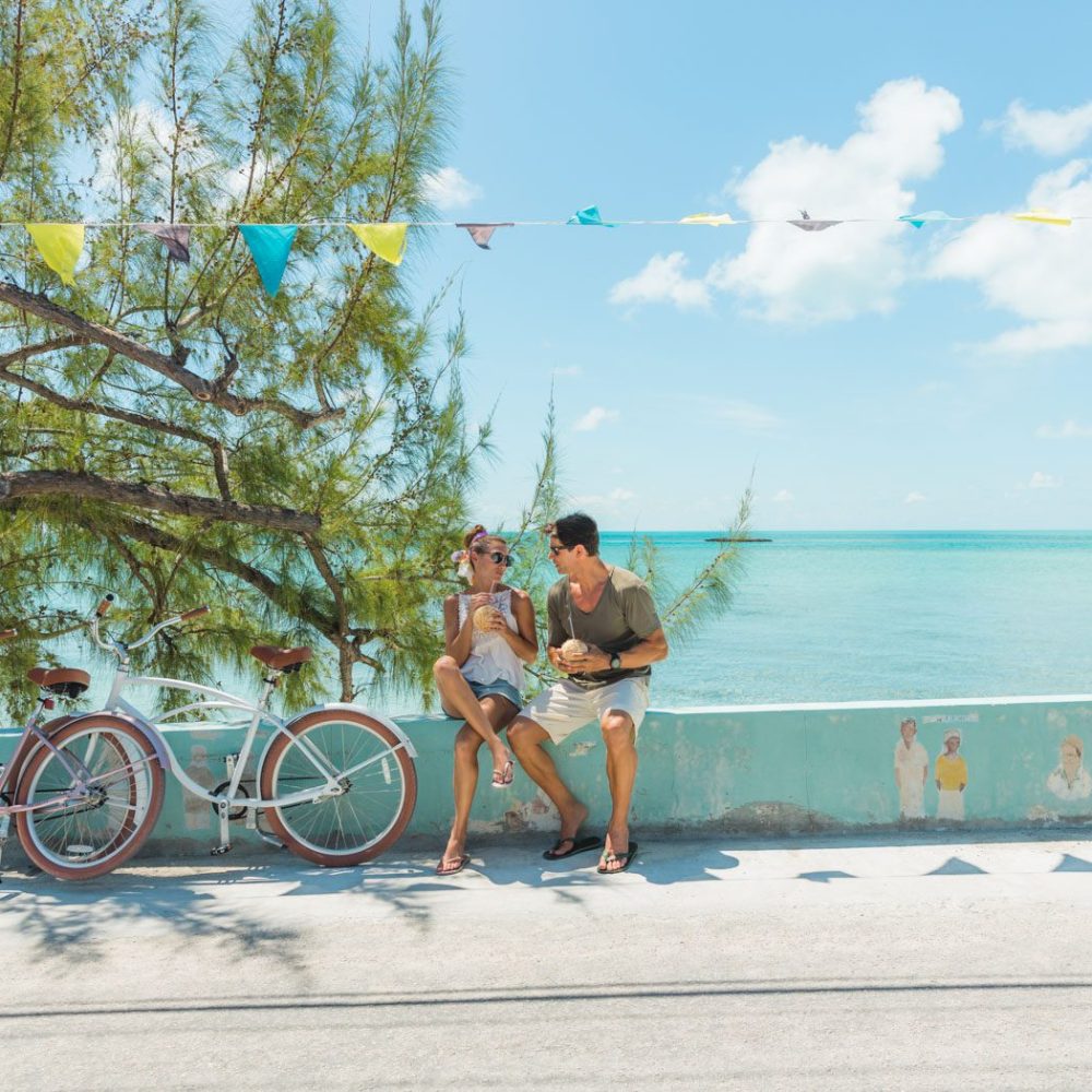 Staniel Cay, Exuma Bahamas Two people sit on a seaside wall, enjoying drinks. Two bicycles are parked nearby. A tree and festive flags hang above, with the ocean in the background.