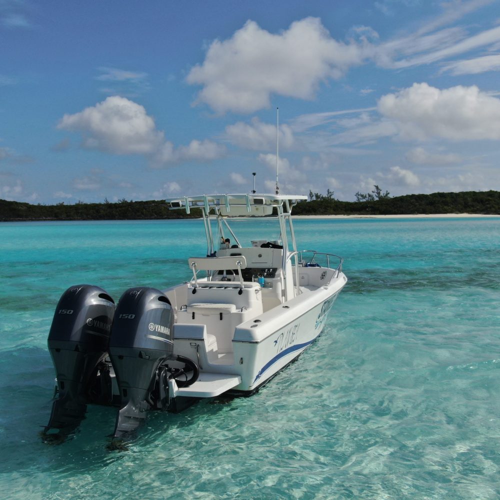 Staniel Cay, Exuma Bahamas A white motorboat with two engines floats on clear turquoise water, surrounded by distant greenery under a partly cloudy sky.