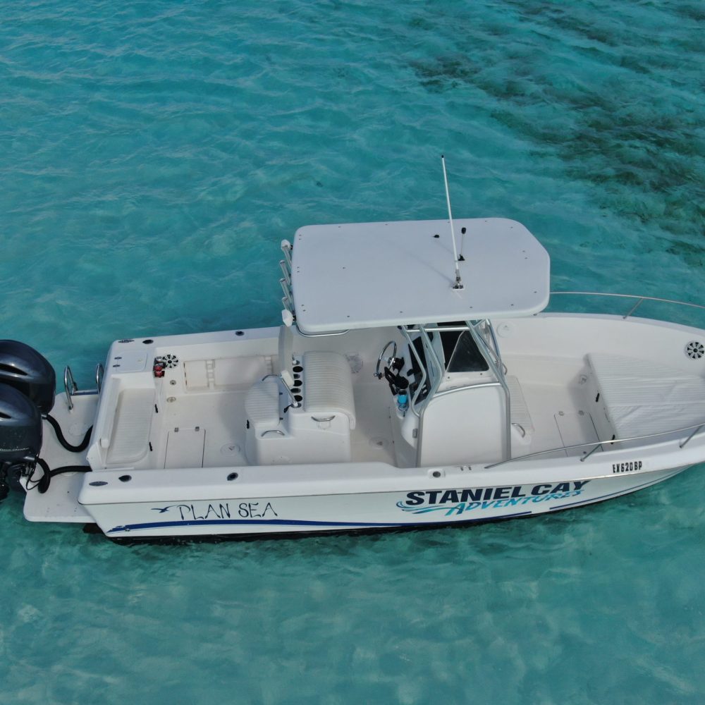 Staniel Cay, Exuma Bahamas A white boat with "Staniel Cay Adventures" written on the side floats on clear, turquoise water. The boat has two outboard motors and seating, with the name "PLAN SEA" visible.