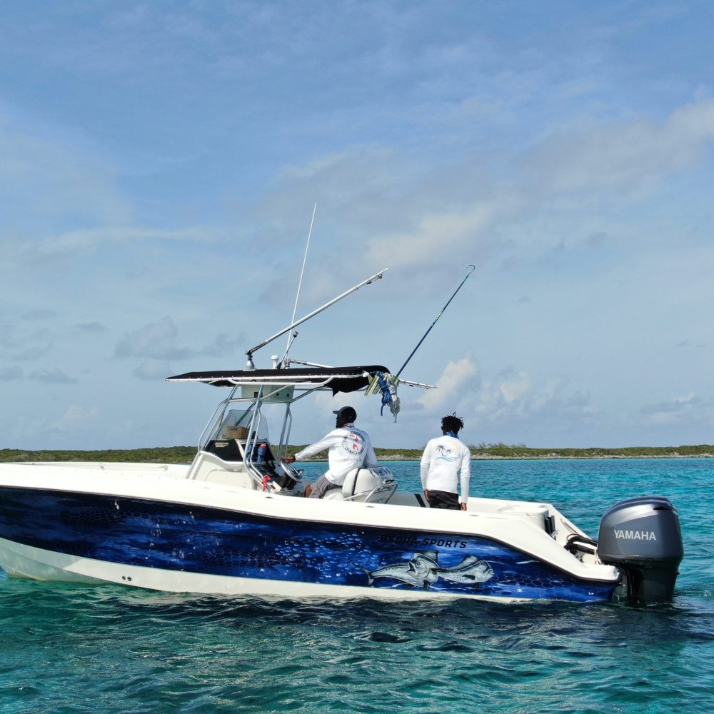 Staniel Cay, Exuma Bahamas A white motorboat with blue graphics on water, two people aboard wearing white shirts and caps, distant land and cloudy sky in the background.