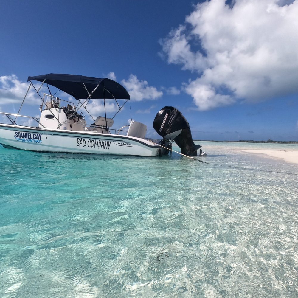 Staniel Cay, Exuma Bahamas A small motorboat with a blue canopy is moored on a clear, shallow beach. The boat's name "Bad Company" and "Staniel Cay" are visible. The sky is partly cloudy.