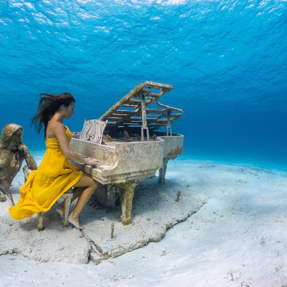 Staniel Cay, Exuma Bahamas A woman in a yellow dress appears to play a submerged piano next to a mermaid statue under the ocean.