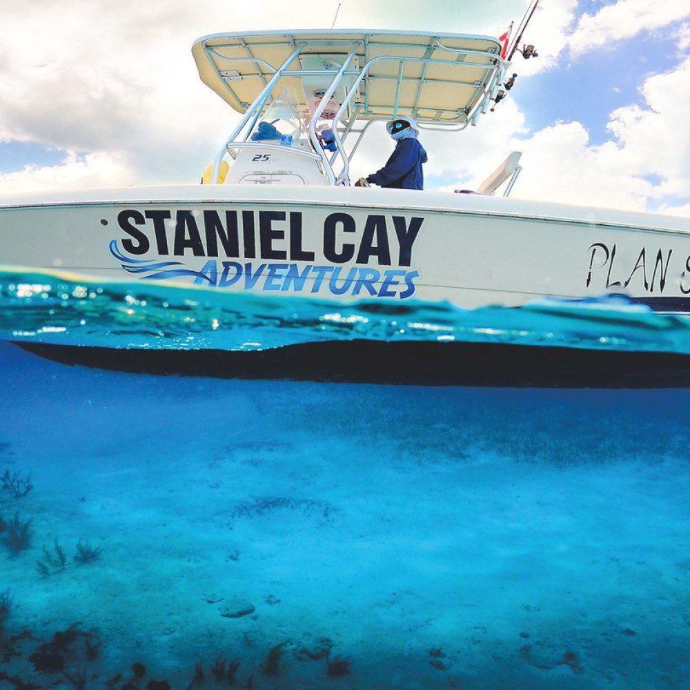 Staniel Cay, Exuma Bahamas Boat labeled "Staniel Cay Adventures" floats on clear blue water, with an underwater view showing coral reef and sea grass beneath.