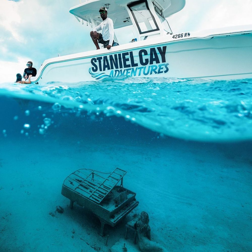 Staniel Cay, Exuma Bahamas A boat labeled "Staniel Cay Adventures" floats on clear water. Underwater, a submerged piano and mermaid statue rest on the sandy seabed.