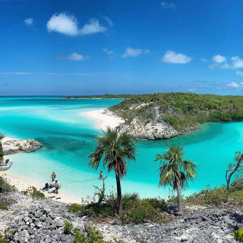 Staniel Cay, Exuma Bahamas Tropical beach scene with turquoise water, white sand, and lush greenery. Several people are relaxing in the sand and shallow water under a blue sky with scattered clouds.