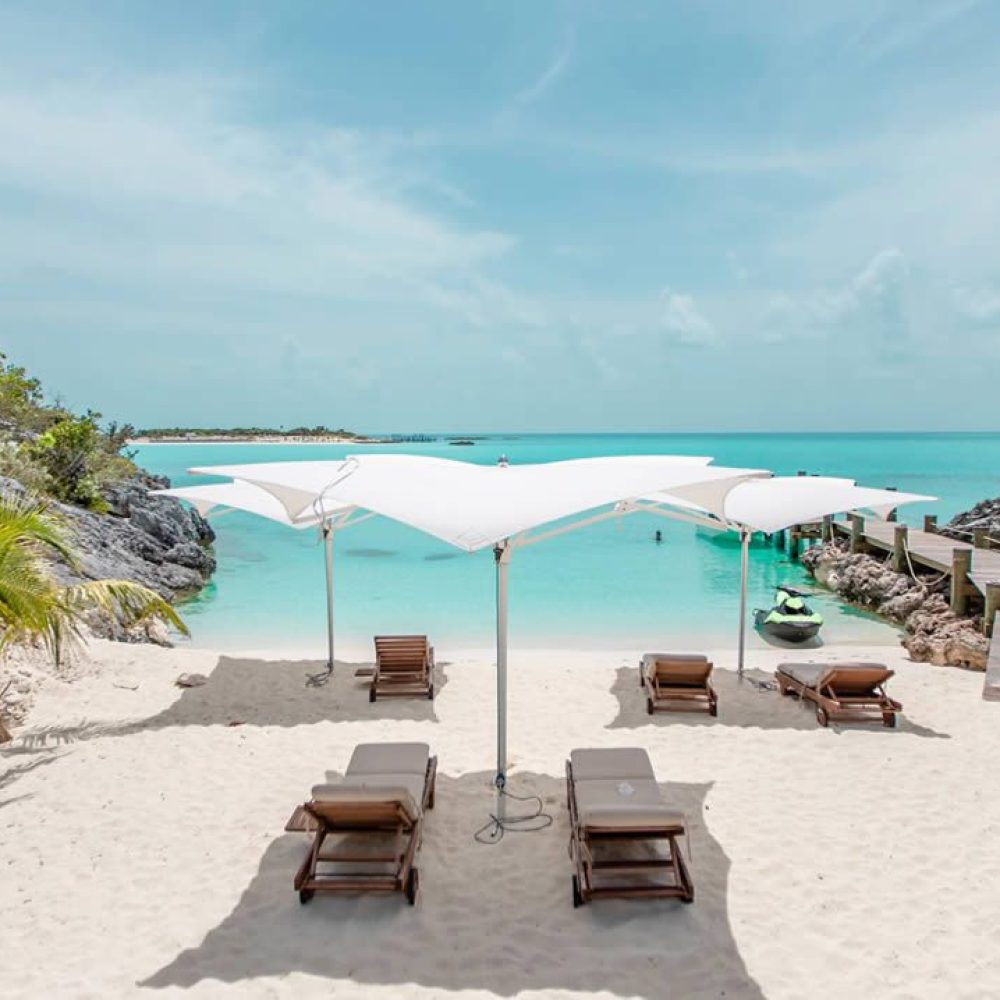 Staniel Cay, Exuma Bahamas Beach scene with six lounge chairs under white canopies facing turquoise water. A wooden pier extends into the sea, surrounded by lush greenery and a clear blue sky.