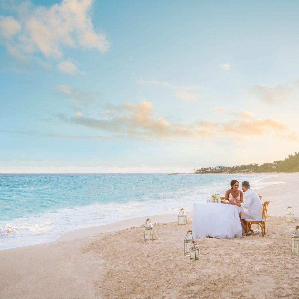 Staniel Cay, Exuma Bahamas A couple sits at a white-clothed table on a sandy beach, surrounded by lanterns, with the ocean and a cloudy sky in the background.