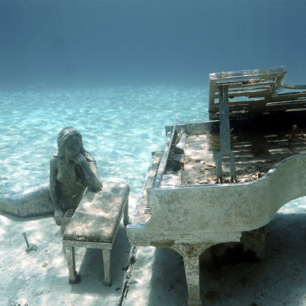 Staniel Cay, Exuma Bahamas Underwater scene featuring a mermaid statue sitting beside a dilapidated piano on a sandy ocean floor.