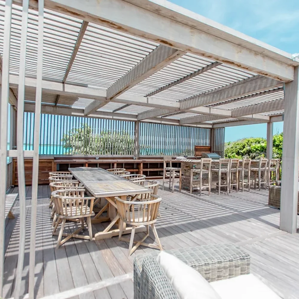 Staniel Cay, Exuma Bahamas Outdoor wooden deck with dining tables and chairs under a pergola, surrounded by greenery and ocean view in the background.