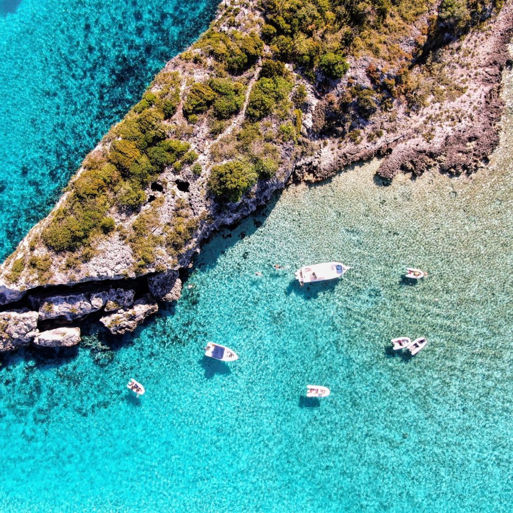 Staniel Cay, Exuma Bahamas Aerial view of boats anchored in clear turquoise water near a rocky, vegetation-covered coastline.