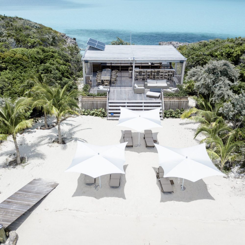 Staniel Cay, Exuma Bahamas Beachfront cabin with a wooden deck, surrounded by greenery. White umbrellas and lounge chairs on the sandy shore. A boardwalk leads to the cabin with a view of turquoise water.