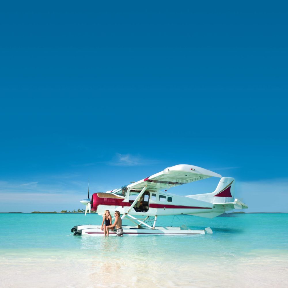 Staniel Cay, Exuma Bahamas A seaplane is anchored in shallow, clear turquoise water near a sandy shore, with one person sitting on the float of the plane under a clear blue sky.