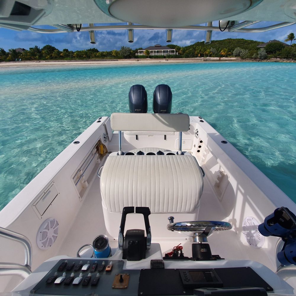 Staniel Cay, Exuma Bahamas View from a small motorboat with seating, facing a clear, shallow turquoise sea. A tropical beach with greenery and a house is visible in the background under a partly cloudy sky.