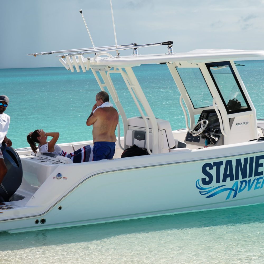 Staniel Cay, Exuma Bahamas A person stands on a white boat with "Staniel Cay Adventures" written on the side, while two others sit and relax in the turquoise ocean.