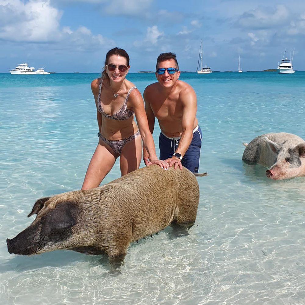 Staniel Cay, Exuma Bahamas A man and woman in swimwear pose with swimming pigs in clear ocean water, with boats visible in the background.